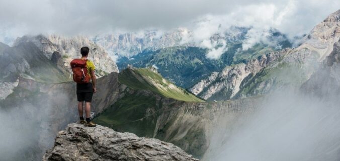 Vaeltaja seisoo kalliojyrkänteellä pilvien peittämien vuorten yllä